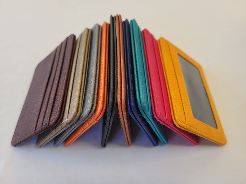 May include: A stack of ten leather card holders in various colors, including brown, orange, gray, blue, teal, and yellow. The card holders are folded in half and arranged in a pyramid shape.