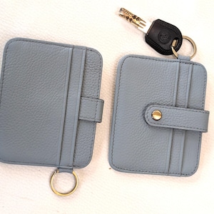 May include: Two light blue leather cardholders with gold-colored key rings. One cardholder has a snap closure, and the other has a tab closure. A key with a black handle is also present.