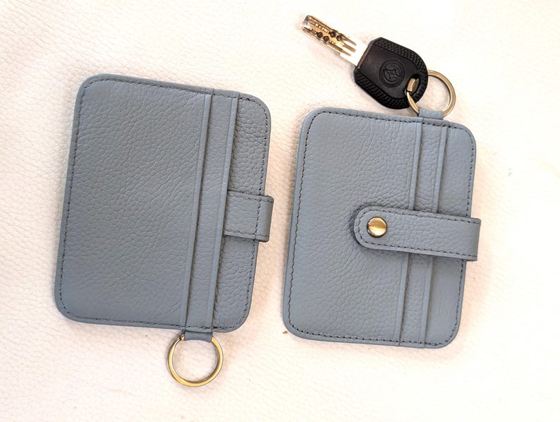 May include: Two light blue leather cardholders with gold-colored key rings. One cardholder has a snap closure, and the other has a tab closure. A key with a black handle is also present.