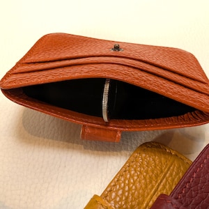May include: An open, orange leather cardholder with a silver coin inside. The cardholder has multiple slots and a small metal clasp. A metal ring is attached to the side. Other leather items in yellow and burgundy are visible.