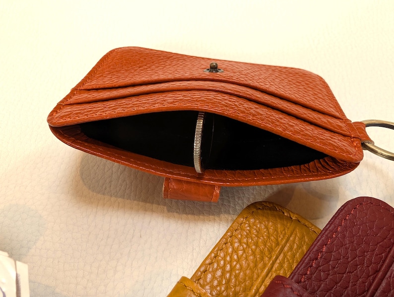 May include: An open, orange leather cardholder with a silver coin inside. The cardholder has multiple slots and a small metal clasp. A metal ring is attached to the side. Other leather items in yellow and burgundy are visible.