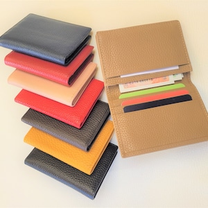 May include: A collection of leather wallets in various colors, including navy, red, tan, and yellow. One wallet is open, displaying cards and cash. The wallets have a textured finish and a classic design, suitable for carrying essentials.