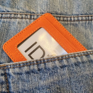May include: An orange leather ID card holder with a clear window for displaying identification. The holder is in a blue denim pocket.