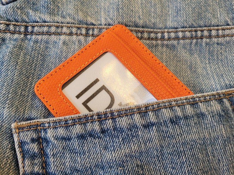 May include: An orange leather ID card holder with a clear window for displaying identification. The holder is in a blue denim pocket.