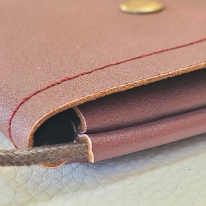 May include: Close-up of a burgundy leather purse with a brown cord strap and a gold button. The purse is folded over and has a red stitch detail.
