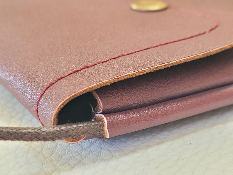 May include: Close-up of a burgundy leather purse with a brown cord strap and a gold button. The purse is folded over and has a red stitch detail.