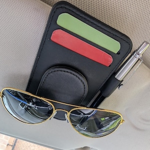 Könnte beinhalten: Ein schwarzer Leder-Visier-Organizer für das Auto mit einem Stifthalter und zwei Kartenfächern. Der Organizer ist an einem Autovisier befestigt und hat eine goldfarbene Sonnenbrille darauf liegen.