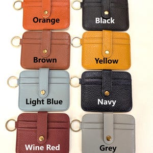 May include: A collection of leather cardholders in various colors, including orange, black, brown, yellow, light blue, navy, wine red, and grey. Each cardholder has a key ring and a snap closure. The text "Orange", "Black", "Brown", "Yellow", "Light Blue", "Navy", "Wine Red", and "Grey" are displayed.