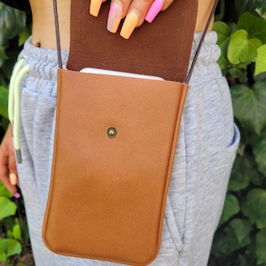 May include: A brown leather crossbody phone bag with a flap closure and a gold button. The bag is worn by a person with colorful nail polish.