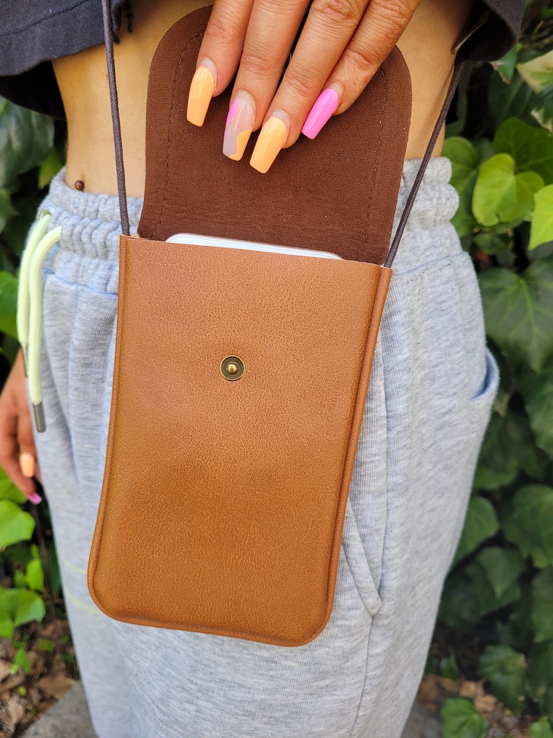 May include: A brown leather crossbody phone bag with a flap closure and a gold button. The bag is worn by a person with colorful nail polish.