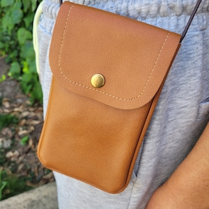 May include: A brown leather crossbody phone bag with a flap closure and a gold button. The bag is worn over a gray sweatshirt.