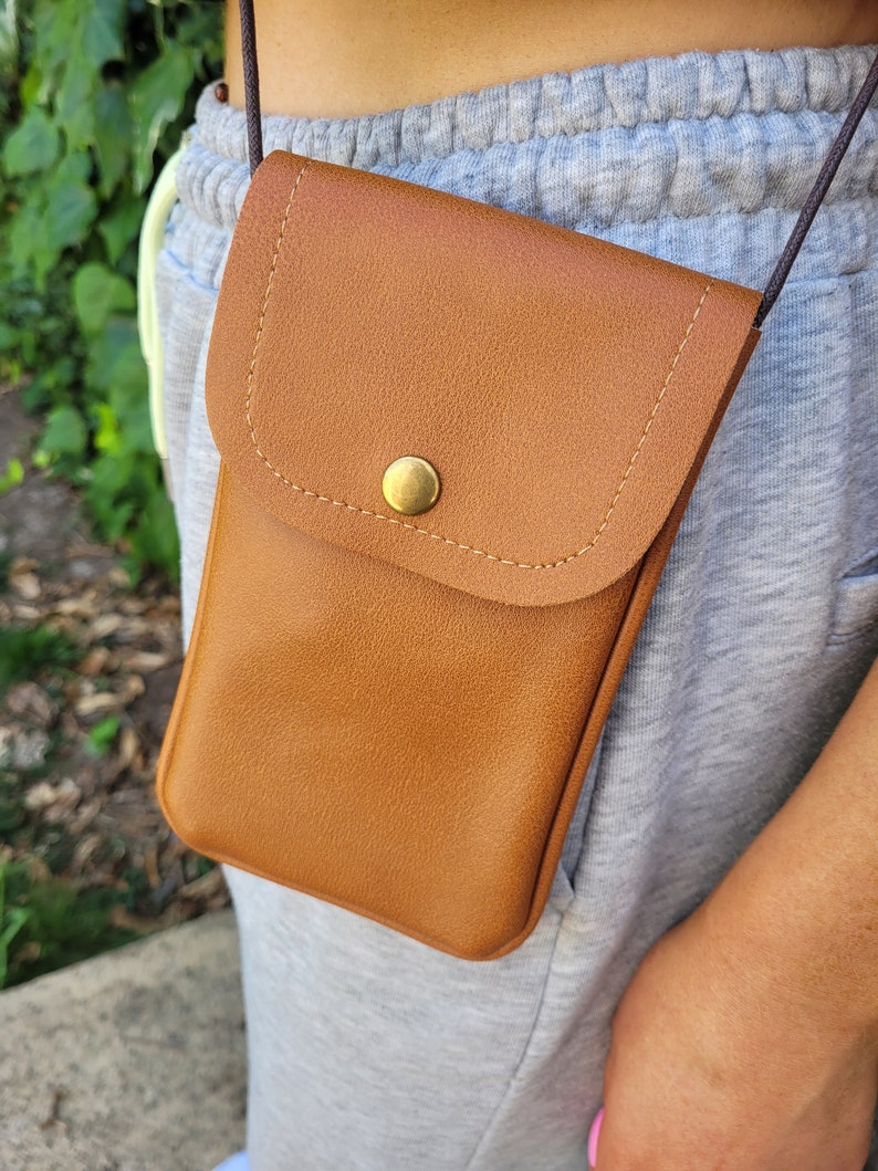 May include: A brown leather crossbody phone bag with a flap closure and a gold button. The bag is worn over a gray sweatshirt.