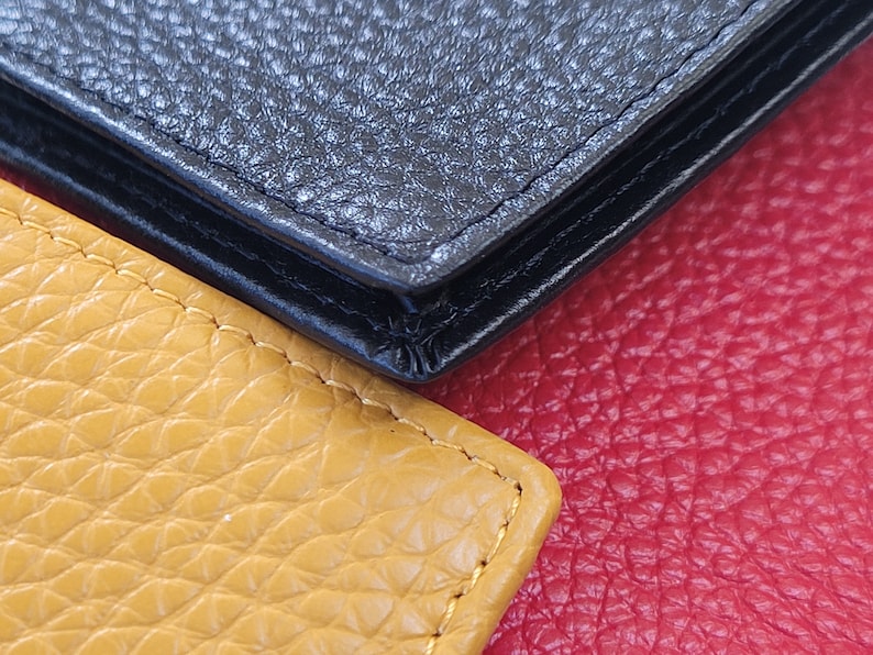 May include: Close-up of three leather textures: black, yellow, and red. The black leather appears to be a wallet or similar item, with visible stitching. The yellow leather is a lighter shade, and the red leather is a deeper tone.