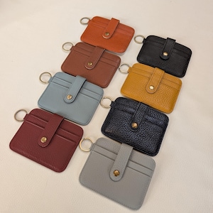 May include: A collection of eight small, rectangular cardholders in various colors, including brown, black, and blue. Each cardholder features a snap closure and a gold-colored key ring. The cardholders are made of a textured material.