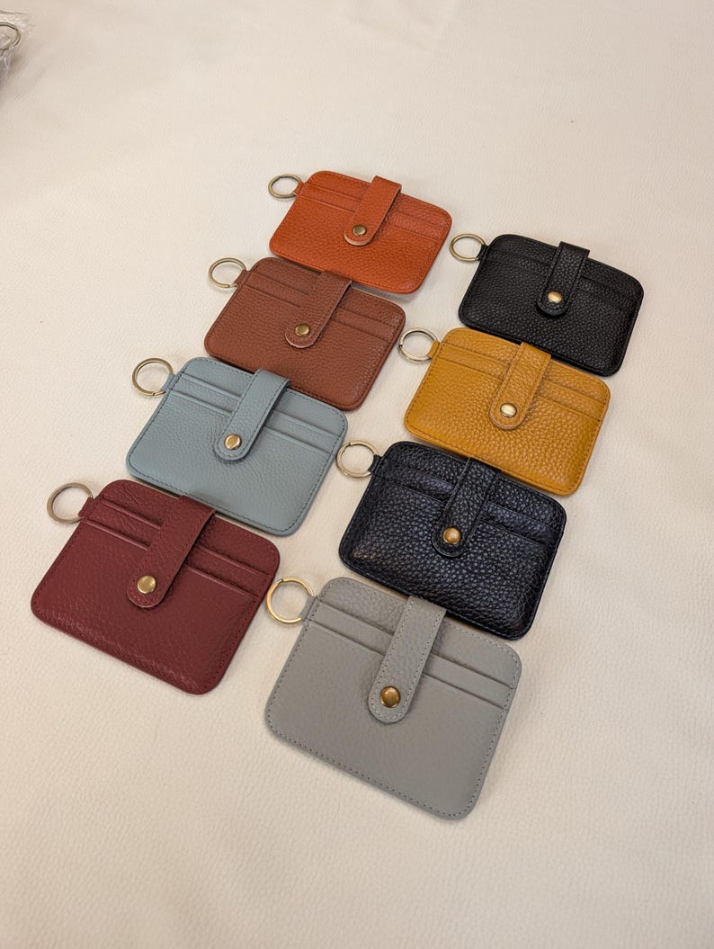 May include: A collection of eight small, rectangular cardholders in various colors, including brown, black, and blue. Each cardholder features a snap closure and a gold-colored key ring. The cardholders are made of a textured material.