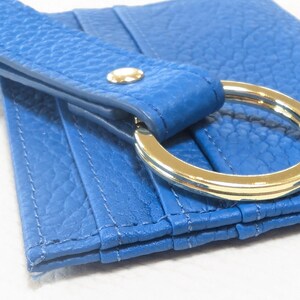 May include: A blue leather card holder with a gold metal ring attached. The card holder has multiple slots for cards and a flap that closes with a snap.