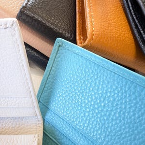May include: A collection of leather wallets in various colors, including white, black, tan, and turquoise. The wallets have a textured surface and visible stitching. The image is a close-up, showcasing the different colors and textures of the wallets.