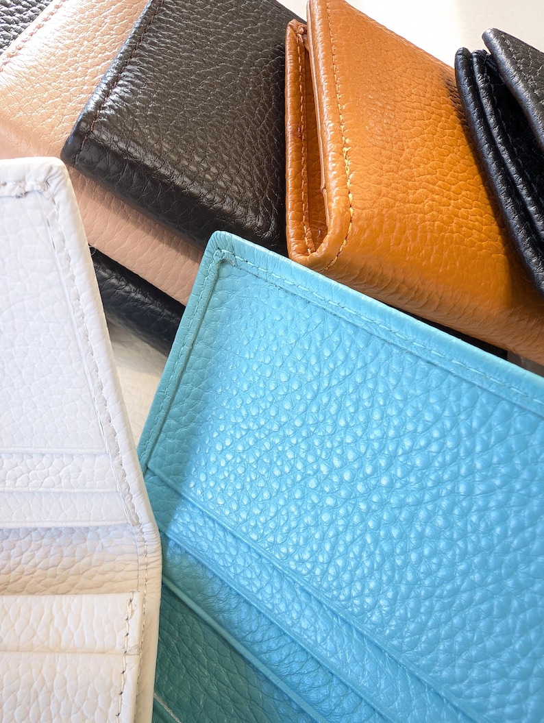 May include: A collection of leather wallets in various colors, including white, black, tan, and turquoise. The wallets have a textured surface and visible stitching. The image is a close-up, showcasing the different colors and textures of the wallets.