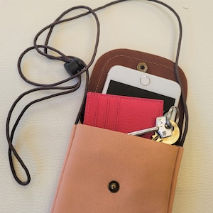 May include: A small, light pink leather crossbody bag with a brown leather flap and a black cord strap. The bag is open and contains a white smartphone, a pink card holder, and a set of keys.