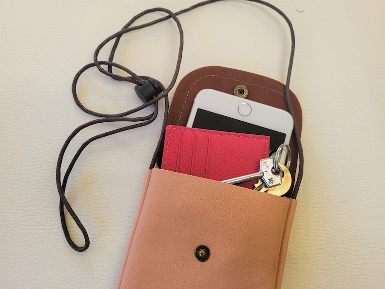 May include: A small, light pink leather crossbody bag with a brown leather flap and a black cord strap. The bag is open and contains a white smartphone, a pink card holder, and a set of keys.
