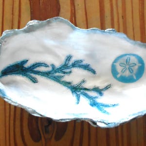 May include: An oyster shell dish with a white interior and a silver edge. It features a teal seaweed design and a teal sand dollar. The dish is displayed on a wooden surface. Measurements are in cm.