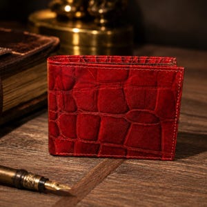 Op de afbeelding: Een felrode bifold portemonnee met een opvallende reliëftextuur van krokodillenleer. De portemonnee heeft een rechthoekige vorm met zichtbare stiksels langs de randen, met een diepe karmozijnrode kleur en een gestructureerd, gevouwen ontwerp.