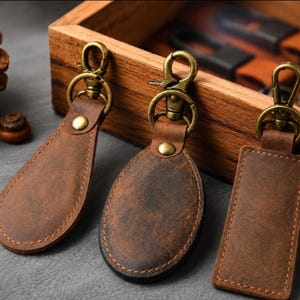 May include: Three brown leather keychains with antique brass-toned metal hardware. Each keychain features a swivel lobster clasp, a circular ring, and a central rivet. The shapes include a teardrop, an oval, and a rectangle, all with visible stitching.