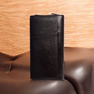 Puede incluir: Una cartera de cuero negro con cierre de cremallera. La cartera es rectangular y tiene una textura suave. Está sobre una superficie de cuero marrón.