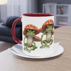 May include: A white ceramic mug with a red rim features two green frogs wearing red and white mushroom hats. The frogs are sitting on a white background.