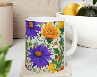 May include: White ceramic mug featuring a vibrant floral design with purple, orange, and yellow flowers. The mug sits on a beige coaster. The background includes a wooden cutting board and a bowl with fruit.