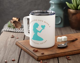 May include: White ceramic travel mug with a teal silhouette of a woman holding a heart. The mug has the text "Embracing Life With A Grateful Heart" printed on it.