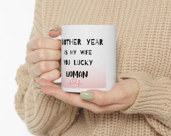 May include: White ceramic mug with black text that reads "Another year as my wife you lucky woman" with a pink and white heart beat graphic.