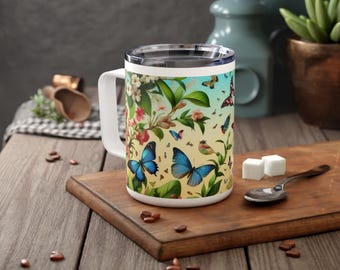 May include: A white ceramic mug with a colorful design featuring butterflies and flowers. The mug has a black handle and a stainless steel lid.