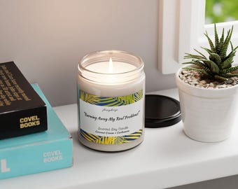 May include: A white soy candle with a yellow and blue label that reads "Burning Away My Real Problems." The candle is in a glass jar with a black lid. The candle is on a white shelf with two books and a potted plant.