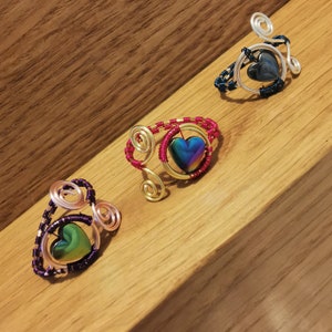 May include: Three wire wrapped rings featuring iridescent heart-shaped stones. The rings are crafted with gold, silver, and copper wire. The stones are set within a wire frame.