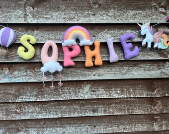 Könnte beinhalten: Ein Filzbanner mit dem Namen "SOPHIE", der in bunten Buchstaben ausgeschrieben ist. Der Banner zeigt außerdem einen Regenbogen, eine Wolke mit Regentropfen, einen Heißluftballon und ein Einhorn.