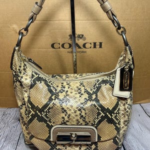 May include: A Coach shoulder bag with a snakeskin pattern in tan, brown, and black. The bag has a curved shape, a zip closure, and a flap with a silver-tone clasp. The Coach logo is visible on the bag and the background.