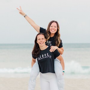 May include: Two women are smiling and laughing on a beach. One woman is giving a peace sign with her hand raised in the air. The woman in the back is giving a piggyback ride to the woman in the front. Both women are wearing black t-shirts with white text that says "LOVE YOURSELF".