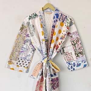 Op de afbeelding: Een witte katoenen badjas met een patchwork-ontwerp met kleurrijke bloemen- en geometrische patronen. De badjas heeft een strikceintuur en lange mouwen.