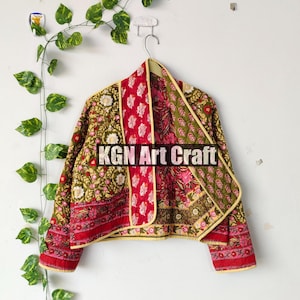Peut inclure: Veste matelassée colorée à motifs floraux rouges, verts et blancs. La veste est suspendue à un cintre sur un mur blanc, avec une vigne de feuilles vertes en arrière-plan. Le texte "KGN Art Craft" est visible sur la veste.