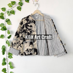 Może przedstawiać: Czarno-biała pikowana kurtka z kwiatowym nadrukiem po jednej stronie i paskowanym wzorem po drugiej. Kurtka ma zapięcie na guziki i kieszeń z przodu. Kurtka wisi na drewnianym wieszaku na tle białej ściany.