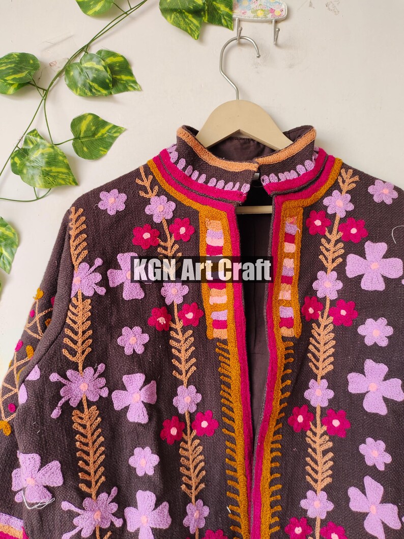 Puede incluir: Una chaqueta marr&oacute;n bordada con dise&ntilde;os florales de color rosa, naranja y rojo. La chaqueta tiene un cuello alto y una cremallera frontal. El texto "KGN Art Craft" es visible en la parte delantera de la chaqueta.