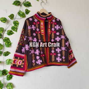 Puede incluir: Una chaqueta marr&oacute;n bordada con dise&ntilde;os florales de color rosa, naranja y blanco. La chaqueta tiene un cuello alto y cierre de bot&oacute;n. El texto "KGN Art Craft" es visible en la parte delantera de la chaqueta.