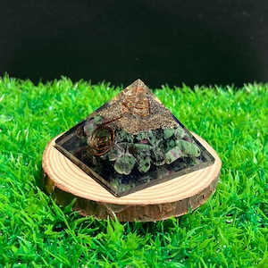 May include: A green and pink crystal pyramid with a copper wire spiral inside. The pyramid is sitting on a wooden base and is surrounded by green grass.