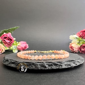 May include: A peach-coloured gemstone bead bracelet with gold-coloured metal detailing. The bracelet is displayed on a black, textured surface, with pink roses and greenery in the background. The text "Gemstone gallery" is visible on the surface.