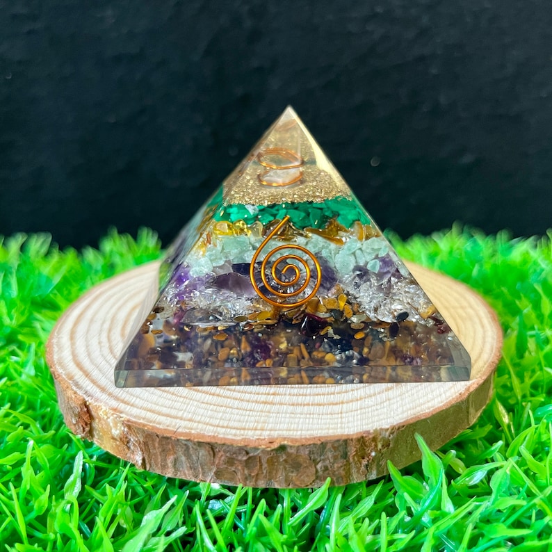 May include: A clear crystal pyramid with seven layers of different coloured crystals, including amethyst, clear quartz, tiger's eye, green aventurine, and malachite. The pyramid is sitting on a wooden base.