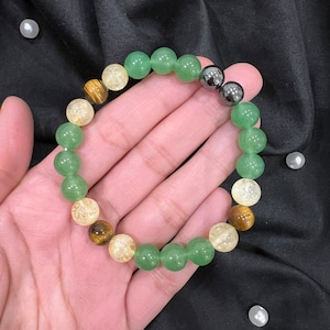 Peut inclure: Un bracelet en perles vertes, jaunes et brunes avec des accents métalliques noirs. Le bracelet est fait de pierres naturelles et est porté au poignet.