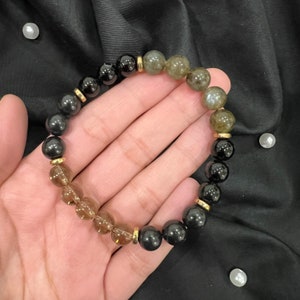May include: A bracelet made of black, brown, and green gemstone beads with gold accents. The bracelet is worn on a person's wrist.