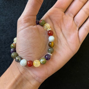 May include: A bracelet made with various colored gemstone beads, including green, brown, red, purple, and white. The bracelet is strung on a silver chain.