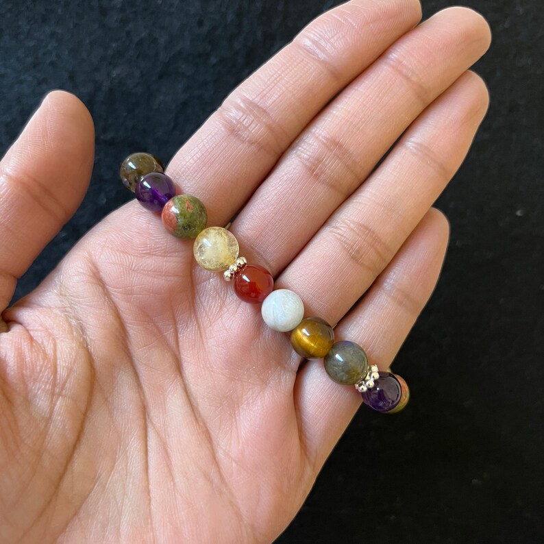 May include: A bracelet made of various colored gemstone beads, including amethyst, green, gold, red, white, brown, and gray. The beads are separated by small gold spacers.